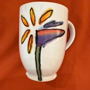 May include: White ceramic mug with a hand-painted design of a stylized flower with orange, yellow, green, purple, and red accents.