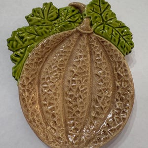 May include: A ceramic cantaloupe-shaped plate with a textured surface and a light brown color. The plate is adorned with green leaves at the top. This decorative plate is ideal for serving or displaying food.