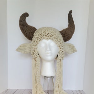 May include: A crocheted white and brown cow hat with long white braids. The hat has large brown horns and small white ears.