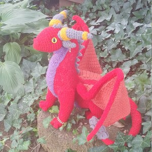 May include: A crocheted red dragon with yellow and grey horns, purple accents, and a red and orange tail. The dragon is sitting on a tree stump.