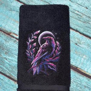 Op de afbeelding: Zwarte handdoek met een geborduurd ontwerp van een raaf voor een maan. De raaf is gedetailleerd met paarse en blauwe tinten, omringd door een bladerkrans. De handdoek is gemaakt van een zacht, absorberend materiaal.