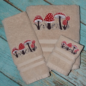 May include: Set of four beige towels with embroidered red and white mushrooms. The towels are different sizes, with the largest towel having three mushrooms and the smallest towel having two mushrooms.