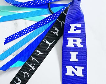 May include: Blue and white hair bow with a blue ribbon that says "ERIN" in white letters. The bow has a black ribbon with white silhouettes of gymnasts.