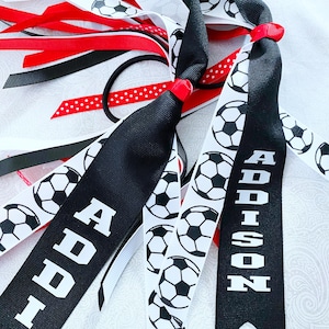 May include: A hair accessory featuring multiple ribbons in black, white, and red. Some ribbons are printed with black and white soccer ball patterns, while others are solid red or white with red polka dots. Two black ribbons feature the name Addison.