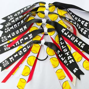 May include: A collection of softball-themed hair ribbons. The ribbons feature a repeating pattern of yellow softballs on a white background. Some ribbons are black with white text, others are red. The text includes names and the word "ALLSTAR".