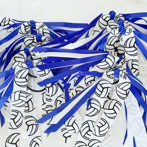 May include: A collection of hair ties featuring multiple ribbons in solid royal blue and white, interspersed with white ribbons printed with a repeating black volleyball graphic. Each hair accessory is constructed with knotted fabric ribbons.