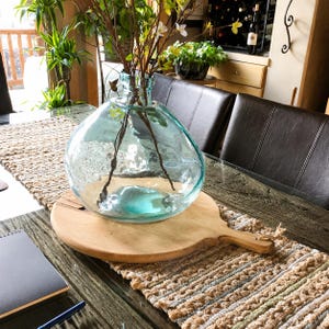 May include: A large, round, light blue glass vase with a thick, clear rim and a slightly irregular, organic shape. The vase sits on a circular wooden serving board with a handle, containing thin, bare brown branches with small yellow flowers.
