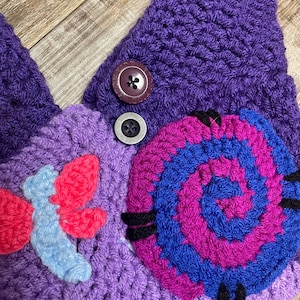 May include: A handmade crochet hat in shades of purple, featuring a spiral design in blue and magenta, and a floral accent in red and light blue. The hat has two buttons and is made of a soft, textured yarn.