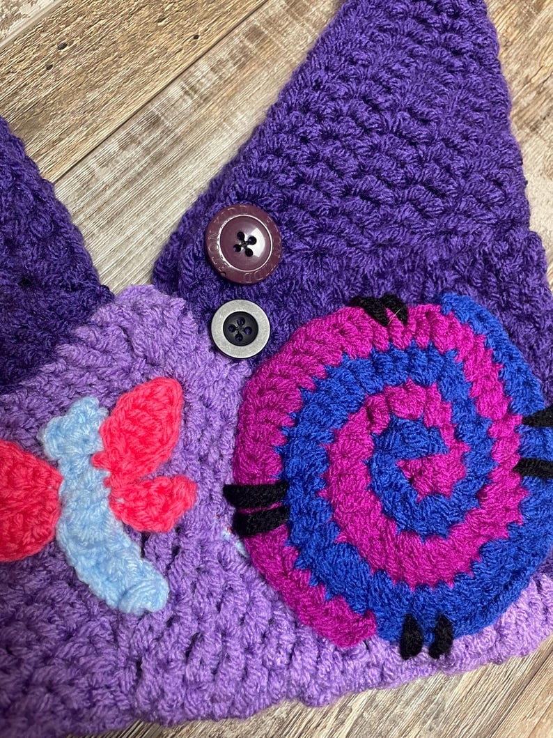 May include: A handmade crochet hat in shades of purple, featuring a spiral design in blue and magenta, and a floral accent in red and light blue. The hat has two buttons and is made of a soft, textured yarn.