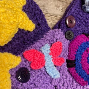 May include: Close-up of a handmade crochet item with a vibrant color palette. The piece features a purple base adorned with yellow stars, a pink and blue butterfly, and a multi-colored spiral design. Dark buttons secure the design.