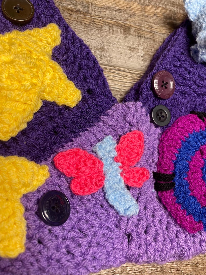 May include: Close-up of a handmade crochet item with a vibrant color palette. The piece features a purple base adorned with yellow stars, a pink and blue butterfly, and a multi-colored spiral design. Dark buttons secure the design.