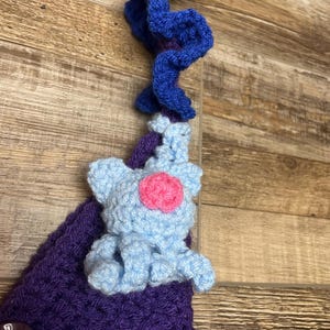 May include: A handmade crochet item featuring a light blue elephant with a pink center, attached to a purple crocheted base. A blue, curly crochet element is at the top. The item is set against a wooden background.
