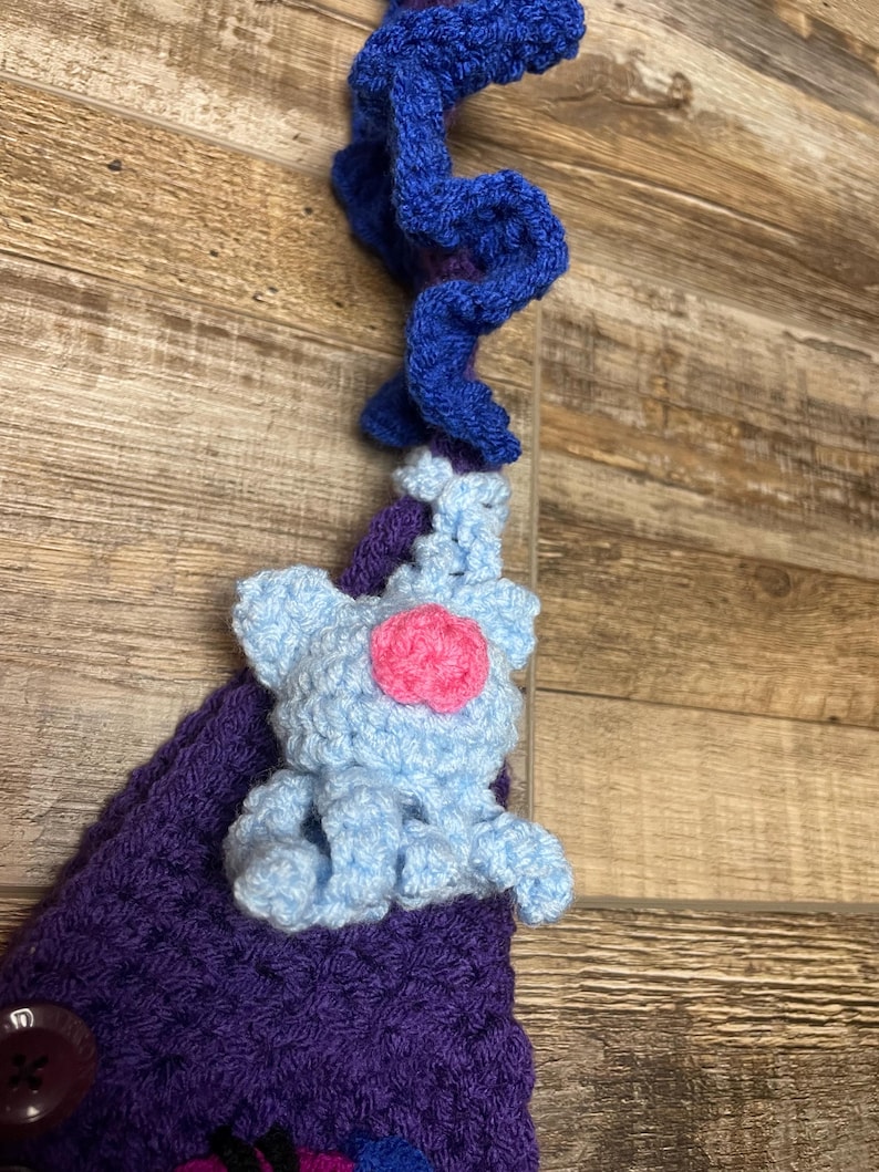 May include: A handmade crochet item featuring a light blue elephant with a pink center, attached to a purple crocheted base. A blue, curly crochet element is at the top. The item is set against a wooden background.