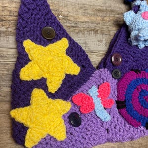 May include: A handmade crochet accessory in shades of purple, yellow, pink, and blue. The piece features yellow star appliqu&eacute;s, a butterfly, and a spiral design, all secured with buttons. The item is likely a whimsical hat or decorative piece.