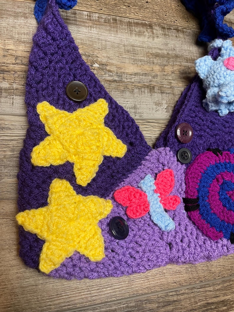 May include: A handmade crochet accessory in shades of purple, yellow, pink, and blue. The piece features yellow star appliqu&eacute;s, a butterfly, and a spiral design, all secured with buttons. The item is likely a whimsical hat or decorative piece.