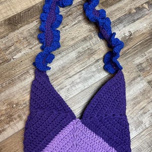 May include: A handmade crochet bag in shades of purple and blue. The bag features a triangular shape with a color-blocked design. The strap is a ruffled design in blue and purple.