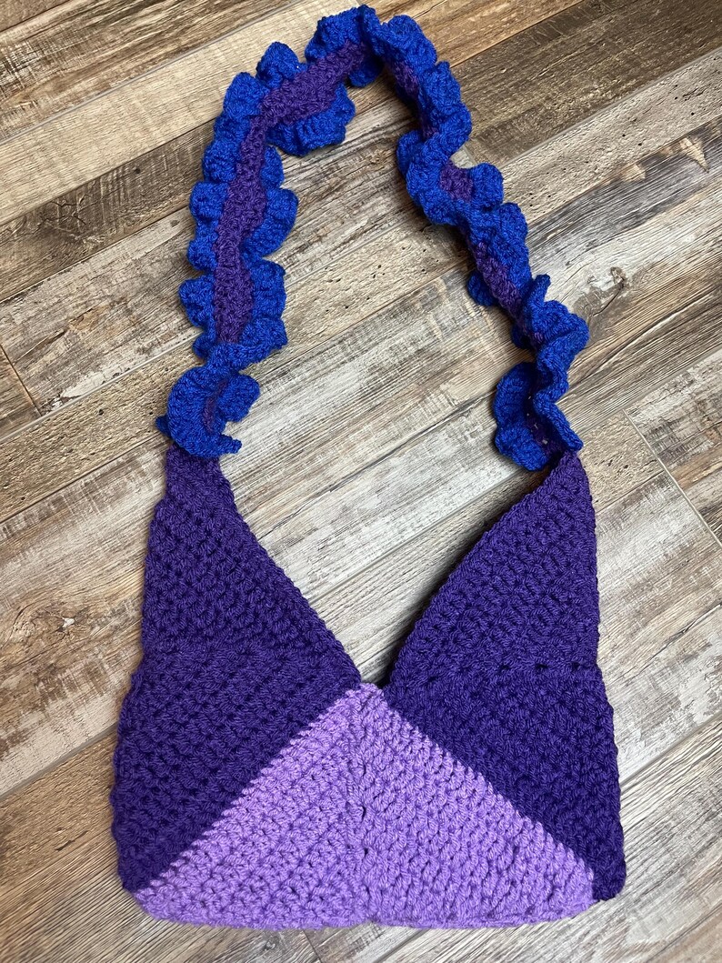 May include: A handmade crochet bag in shades of purple and blue. The bag features a triangular shape with a color-blocked design. The strap is a ruffled design in blue and purple.