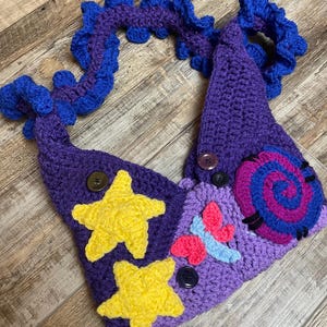 May include: A handmade crochet headpiece in shades of purple and blue, adorned with yellow stars, a pink and blue butterfly, and a spiral design. The headpiece features button accents and a decorative, ruffled edge.