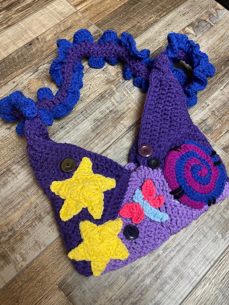 May include: A handmade crochet headpiece in shades of purple and blue, adorned with yellow stars, a pink and blue butterfly, and a spiral design. The headpiece features button accents and a decorative, ruffled edge.