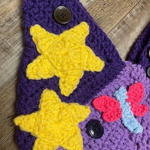 May include: A handmade crochet item in shades of purple, featuring two yellow stars and a pink and blue butterfly. The item is adorned with dark brown buttons. The texture of the crochet is visible, showcasing the craftsmanship.