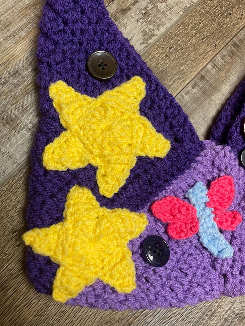 May include: A handmade crochet item in shades of purple, featuring two yellow stars and a pink and blue butterfly. The item is adorned with dark brown buttons. The texture of the crochet is visible, showcasing the craftsmanship.