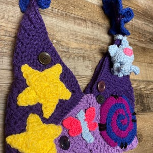 May include: A handmade crochet bag in shades of purple, adorned with yellow stars, a pink and blue butterfly, and a blue and purple spiral. The bag features a blue strap and a small blue creature with a pink heart.