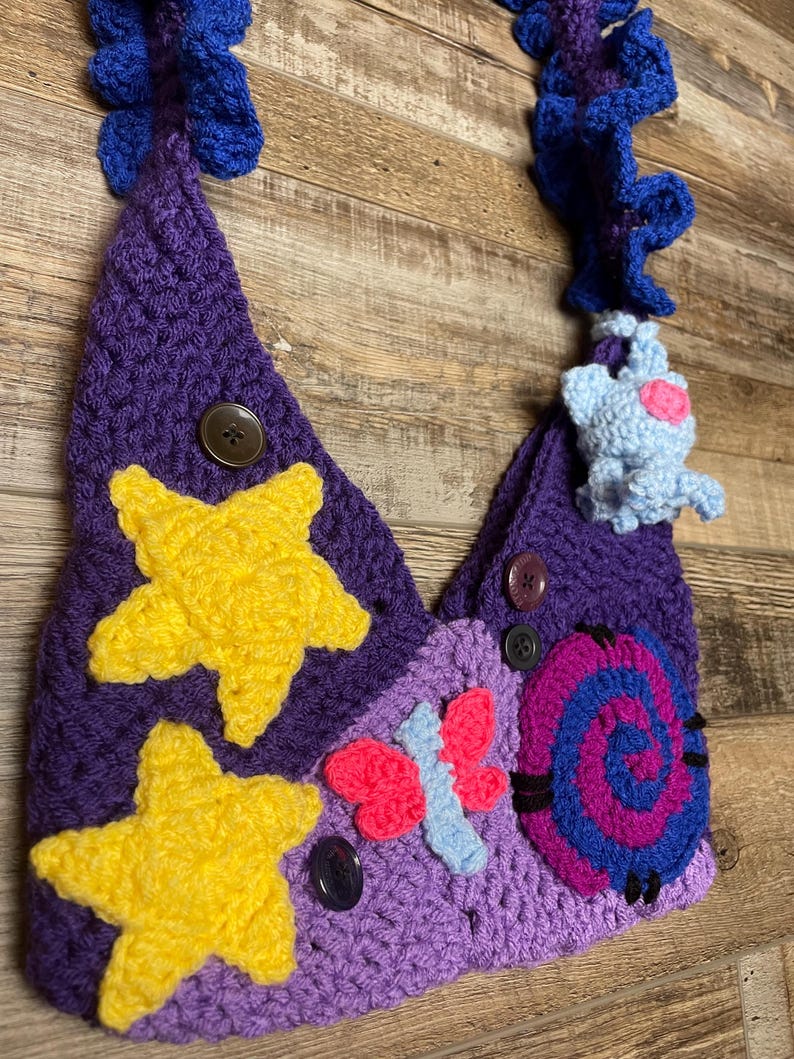 May include: A handmade crochet bag in shades of purple, adorned with yellow stars, a pink and blue butterfly, and a blue and purple spiral. The bag features a blue strap and a small blue creature with a pink heart.
