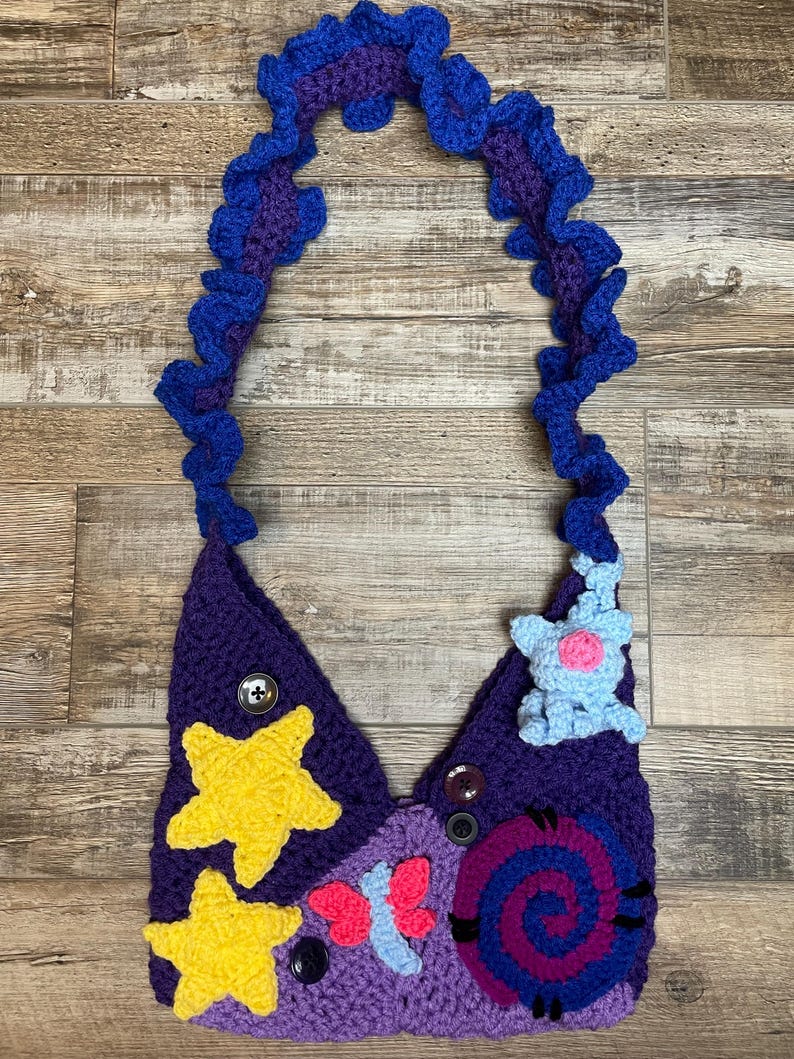 May include: A handmade crochet bag in shades of purple, blue, and yellow. The bag features a whimsical design with yellow stars, a pink and blue butterfly, and a blue creature. The strap is a ruffled blue and purple design.