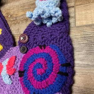 May include: A handmade crochet hat in shades of purple, blue, pink, and yellow. The hat features a spiral design, a blue and pink creature, and a butterfly. Two buttons are sewn on the hat.
