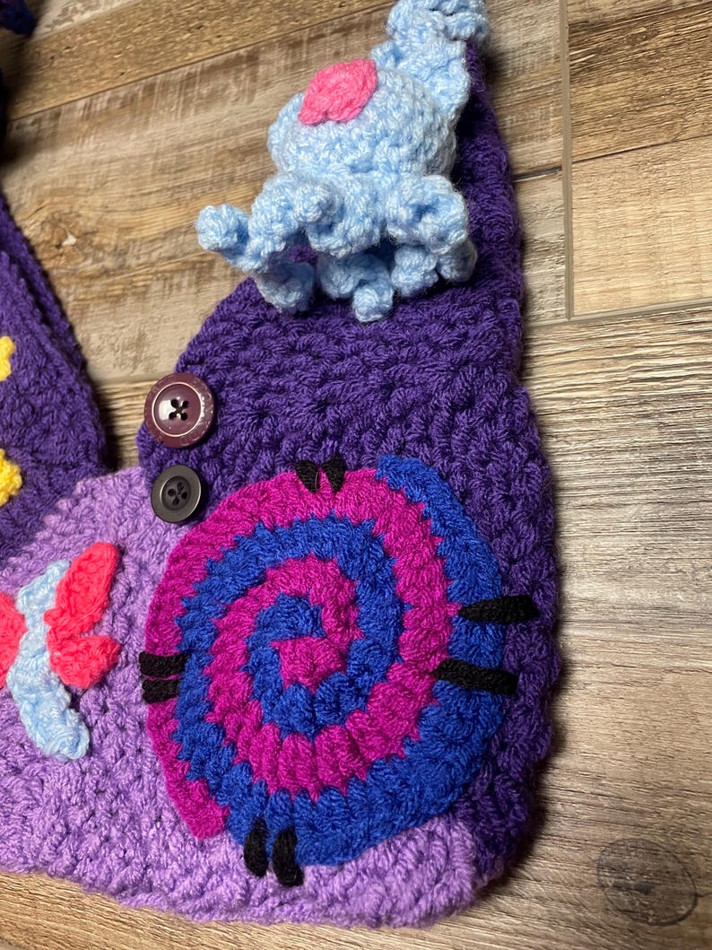 May include: A handmade crochet hat in shades of purple, blue, pink, and yellow. The hat features a spiral design, a blue and pink creature, and a butterfly. Two buttons are sewn on the hat.