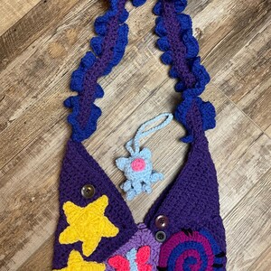 May include: Handmade crochet bag in shades of purple, with yellow star appliqu&eacute;s and a blue and pink spiral design. The strap is a ruffled design in blue and purple. Includes a small blue crochet charm.