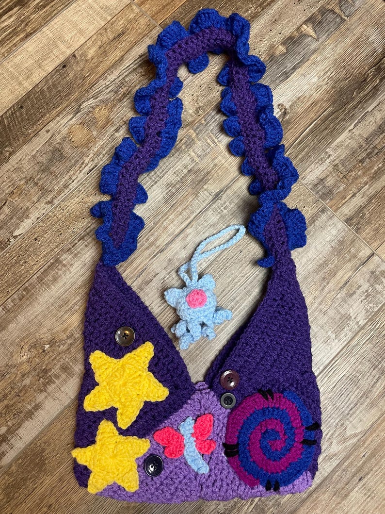 May include: Handmade crochet bag in shades of purple, with yellow star appliqu&eacute;s and a blue and pink spiral design. The strap is a ruffled design in blue and purple. Includes a small blue crochet charm.