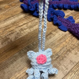 May include: A light blue crochet ornament with a pink center and dangling legs. The ornament is held by a light blue crochet string. Other blue and purple crochet pieces are in the background.