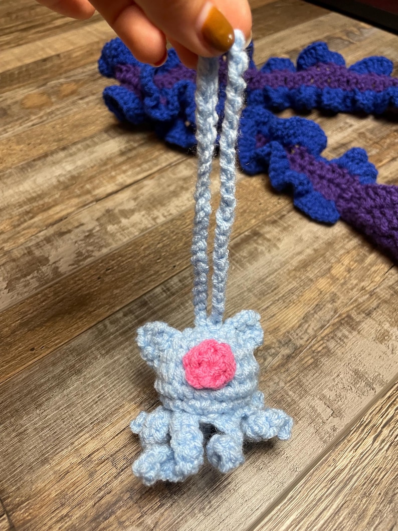 May include: A light blue crochet ornament with a pink center and dangling legs. The ornament is held by a light blue crochet string. Other blue and purple crochet pieces are in the background.