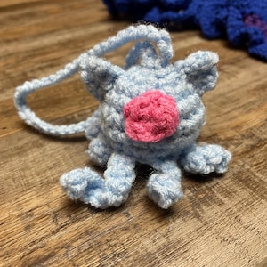 May include: A light blue crocheted creature with a pink nose and small ears. The handmade plush toy has a loop for hanging and is sitting on a wooden surface. Another blue and purple crocheted item is in the background.