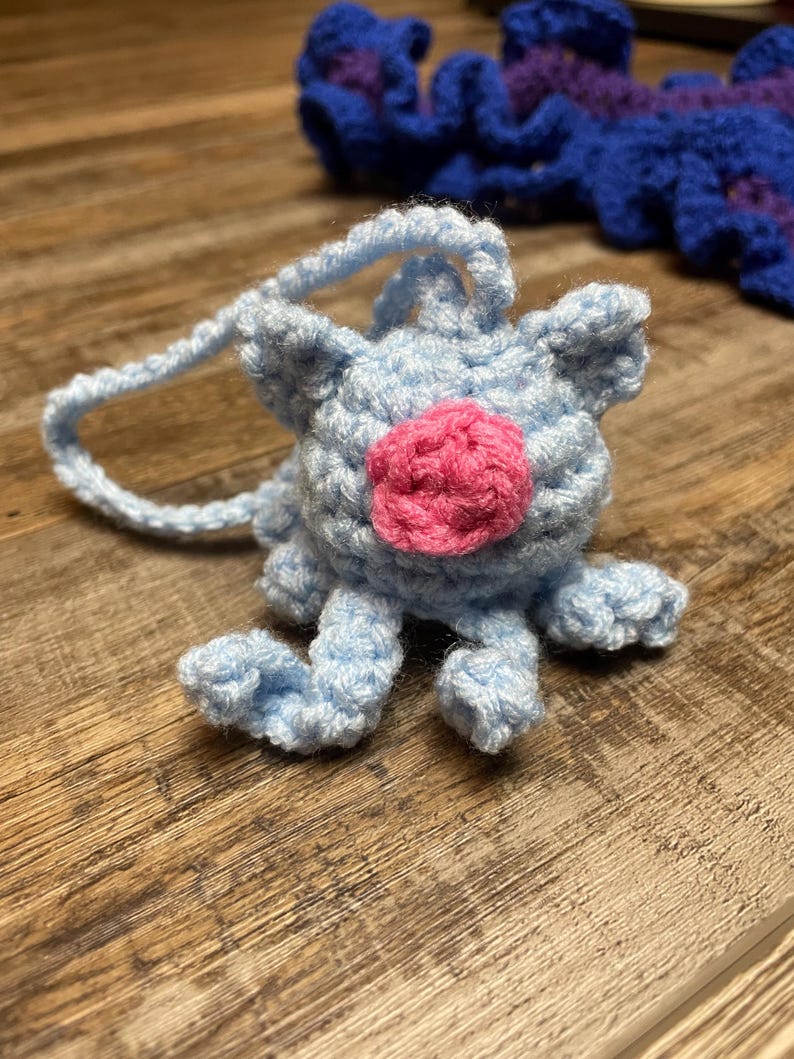 May include: A light blue crocheted creature with a pink nose and small ears. The handmade plush toy has a loop for hanging and is sitting on a wooden surface. Another blue and purple crocheted item is in the background.