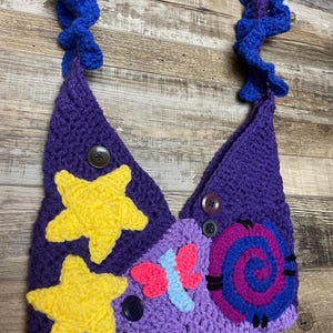 May include: A handmade crochet bag in shades of purple, with yellow star and butterfly accents. The bag features a spiral design and is adorned with buttons. The strap is a ruffled blue and purple design.