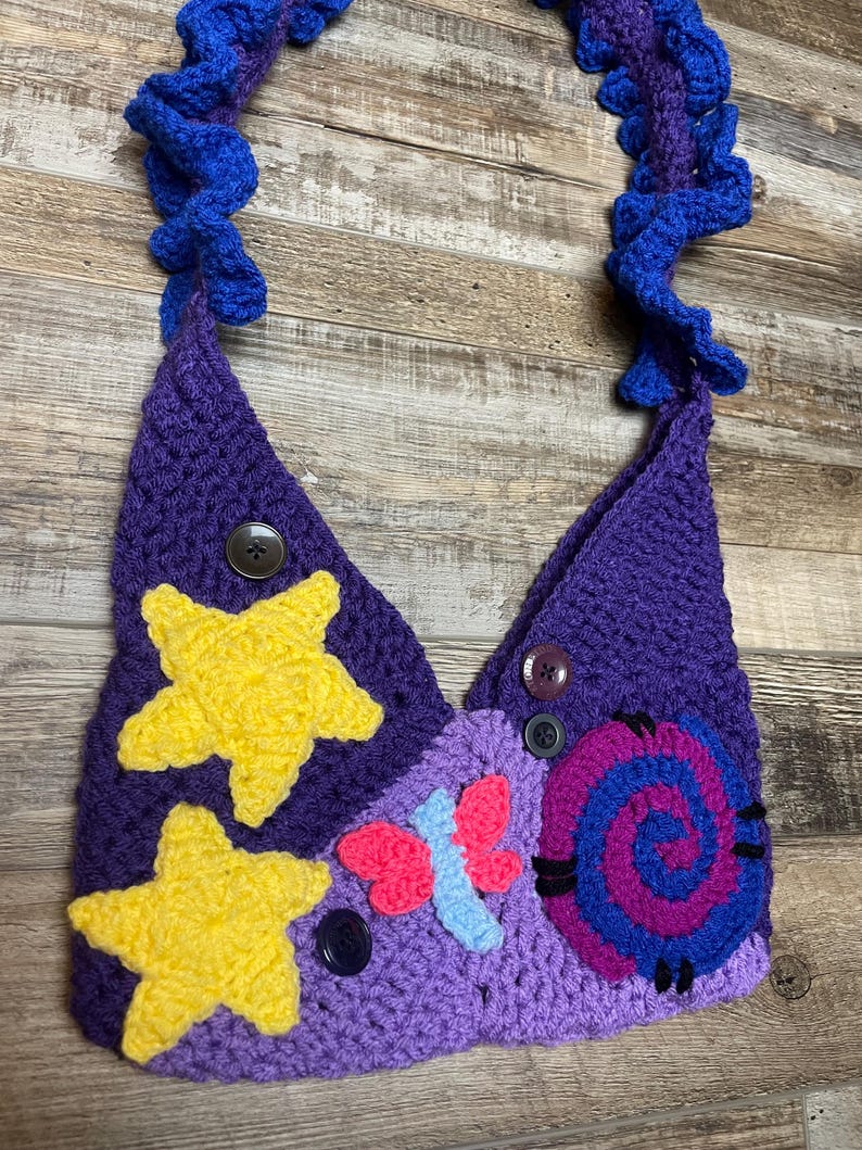 May include: A handmade crochet bag in shades of purple, with yellow star and butterfly accents. The bag features a spiral design and is adorned with buttons. The strap is a ruffled blue and purple design.