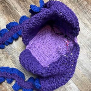 May include: A handmade crochet hat in shades of purple and blue, resembling a dragon. The hat features a rounded crown, a face area, and a tail-like extension with blue accents. The crochet work is visible, showcasing the texture and craftsmanship.