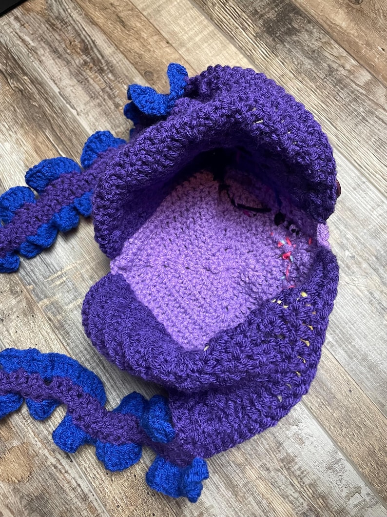 May include: A handmade crochet hat in shades of purple and blue, resembling a dragon. The hat features a rounded crown, a face area, and a tail-like extension with blue accents. The crochet work is visible, showcasing the texture and craftsmanship.