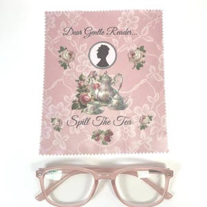 May include: Pink eyeglasses with a rectangular frame sit below a pink cleaning cloth. The cloth features a floral design, a silhouette, and the text "Dear Gentle Reader... Spill The Tea."