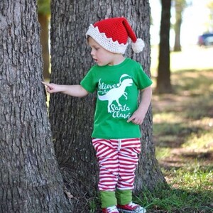 May include: A young child wearing a red and white crocheted Santa hat, a green t-shirt with a white dinosaur graphic and the text "Believe in Santa Claws", red and white striped trousers, and green and white trainers. The child is standing in front of a tree.