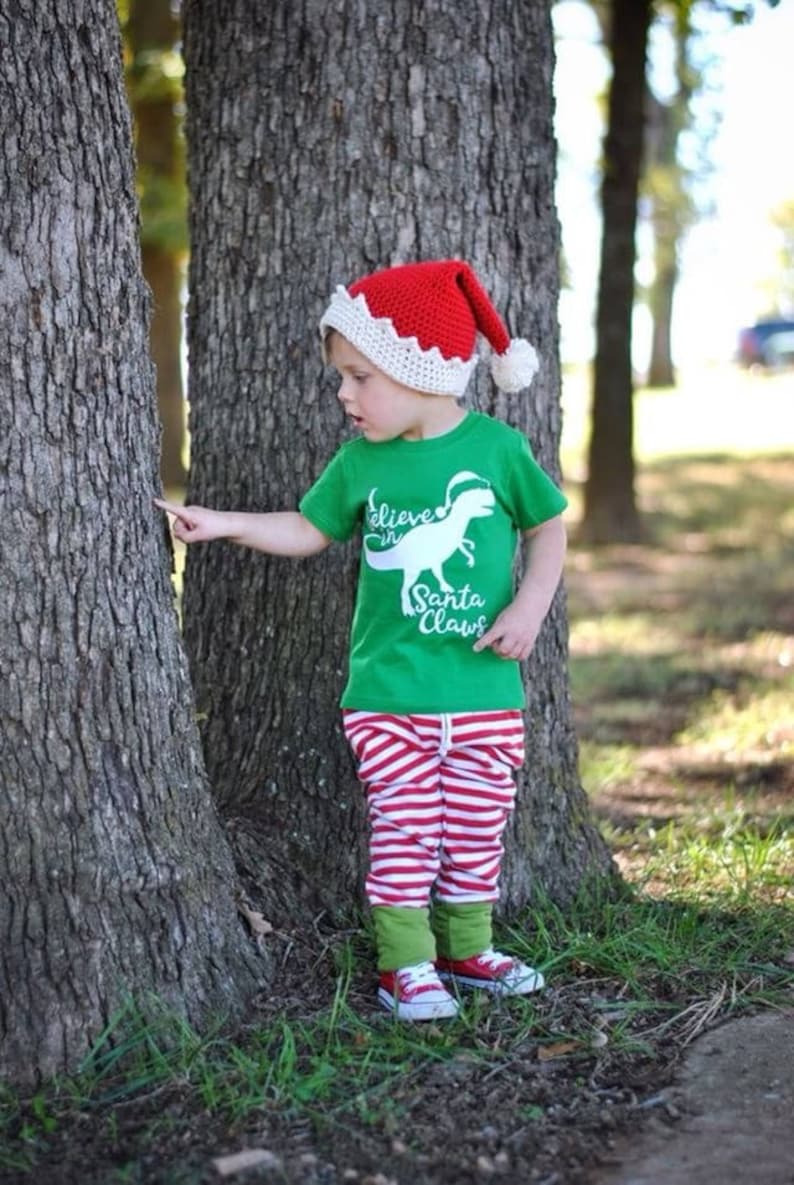 May include: A young child wearing a red and white crocheted Santa hat, a green t-shirt with a white dinosaur graphic and the text "Believe in Santa Claws", red and white striped trousers, and green and white trainers. The child is standing in front of a tree.