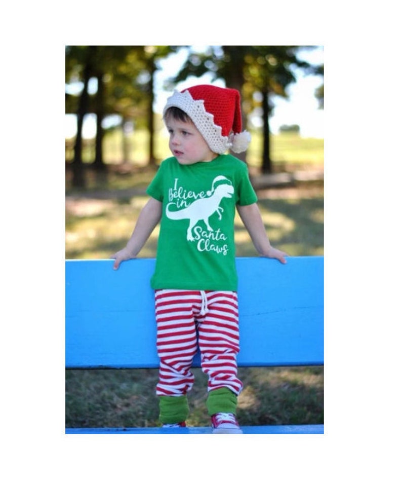 May include: A young child wearing a red and white Santa hat, a green t-shirt with a white dinosaur and the text "I believe in Santa Claws", and red and white striped trousers.