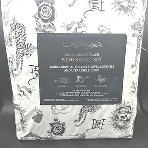 May include: A white king sheet set featuring a black tattoo-style print of tigers, roses, skulls, and hearts. The packaging includes a black label with the text Ed Hardy, Monochrome Flash King Sheet Set, and lists dimensions in inches and centimeters.