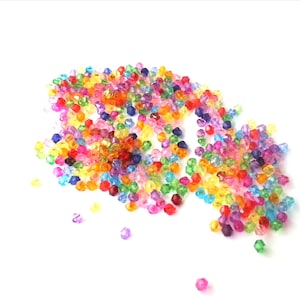 May include: A collection of multi-coloured, faceted beads scattered on a white backdrop. The beads are in shades of red, orange, yellow, green, blue, and purple. These beads are suitable for jewellery making and craft projects.
