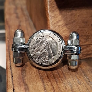 May include: A silver-toned coin spinner with a British 10 pence coin featuring the image of a ship. The coin is mounted on a swiveling mechanism.