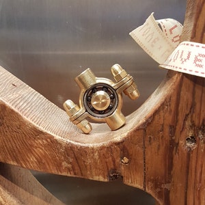 Könnte beinhalten: Ein goldfarbener Metall-Fidget-Spinner mit vier Armen und einem zentralen Lager. Der Spinner liegt auf einem Stück Holz.