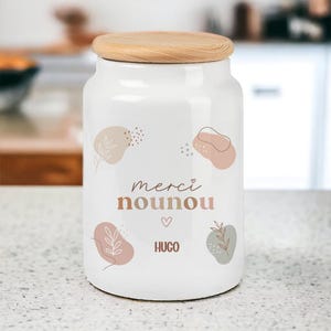 Peut inclure: Un bocal de rangement en céramique blanche de forme cylindrique avec un couvercle en bois clair naturel. L'avant présente le texte "merci nounou" et "HUGO" en lettres marron, entouré de formes abstraites rose et vert avec des feuilles.