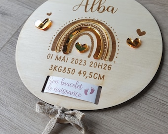May include: A wooden circle with a gold rainbow and the name "Alba" engraved on it. The circle also includes the date "01 MAI 2023 20H26", the weight "3KG850", and the length "49,5CM". There is a small tag attached to the bottom of the circle that says "Son bracelet de naissance".